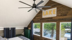 Camera da letto con due letti, una ventola a soffitto, un muro di legno con un cartello 'LAKE' e ampie finestre con vista su un lago.