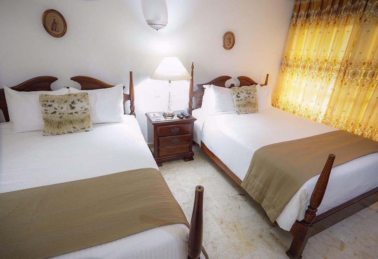 A hotel room with two beds, a nightstand, a lamp, and a window with curtains.