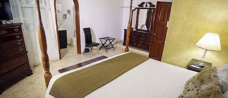 A bedroom with a four-poster bed, a wooden dresser, a chair, a mirror, and a nightstand with a lamp.