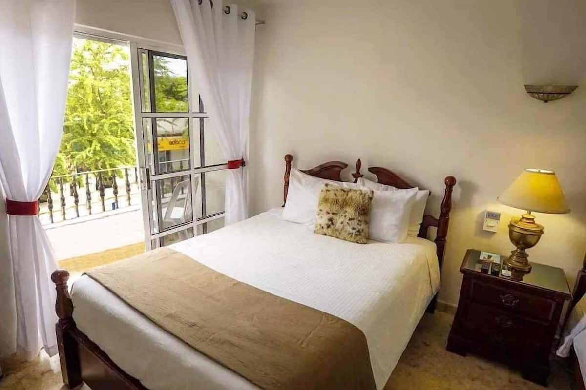 Un dormitorio con cama, mesita de noche, lámpara y ventana con cortinas.