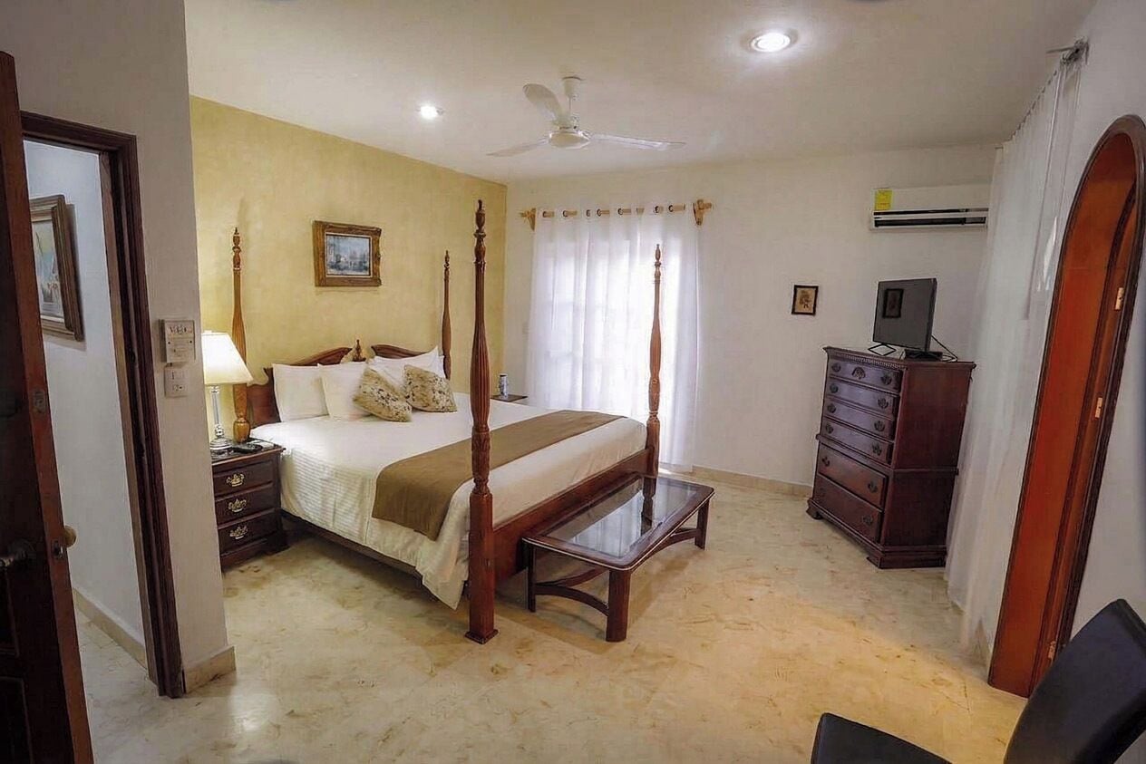 A bedroom with a four-poster bed, a bedside table, a dresser, a ceiling fan, and an air conditioning unit.