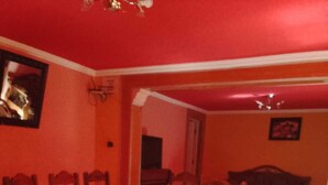 Uma área de refeições com uma mesa e cadeiras de madeira, uma televisão e um sofá ao fundo.