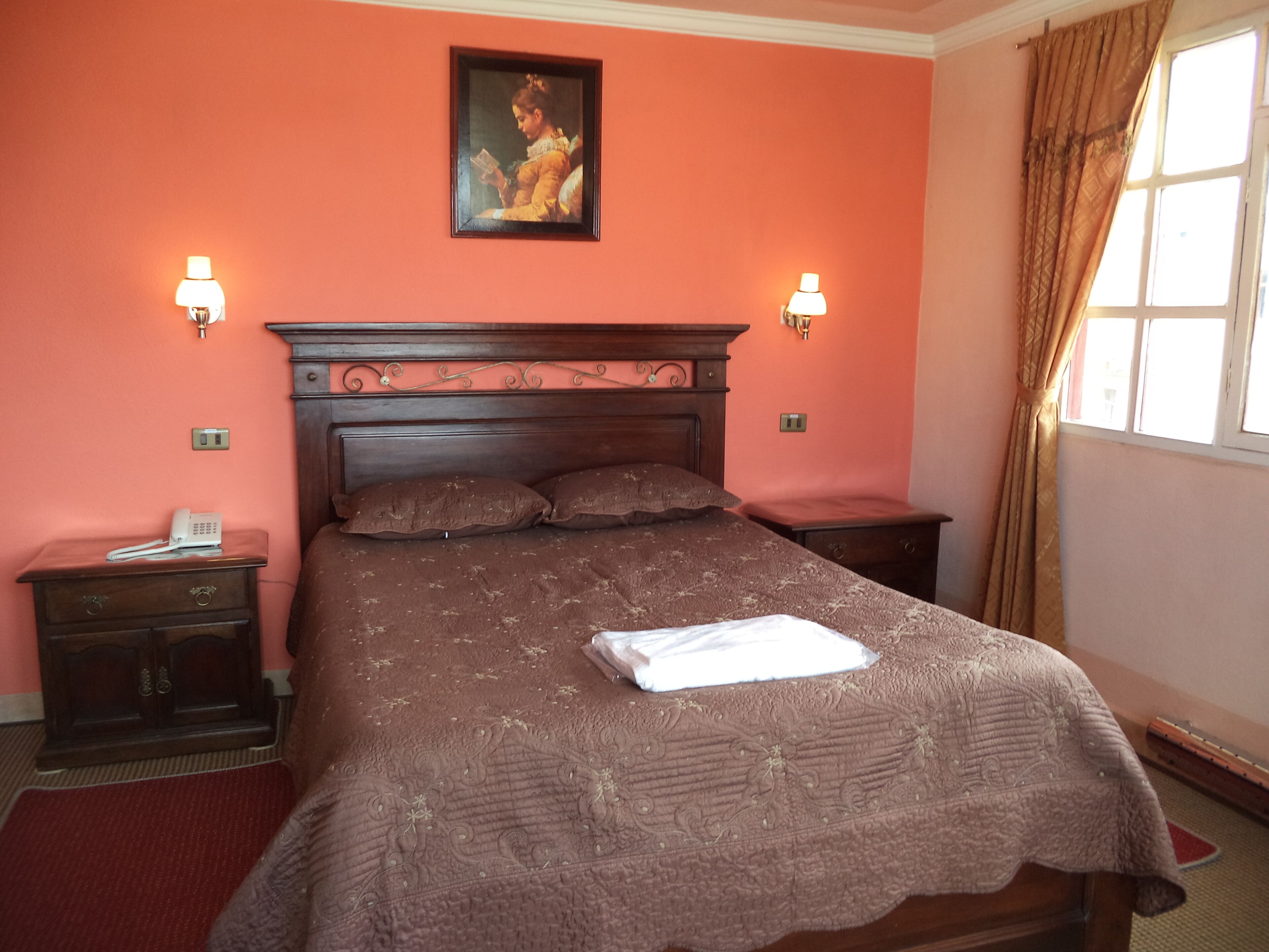 Un dormitorio con cabecera de madera, dos mesitas de noche con lámparas, un cuadro en la pared y una ventana con cortinas.