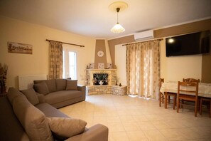 A living room with a sofa, a fireplace, a dining table with chairs, and a television mounted on the wall. - Nikorina Apts | 3 (Apolpena)