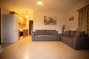A living room with a sofa, a kitchen area with cabinets and appliances, and a painting on the wall. - Nikorina Apts | 3 (Apolpena)