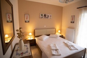 A hotel room with a bed, bedside lamps, a nightstand, a mirror, and a wall with framed artwork. - Nikorina Apts | 3 (Apolpena)