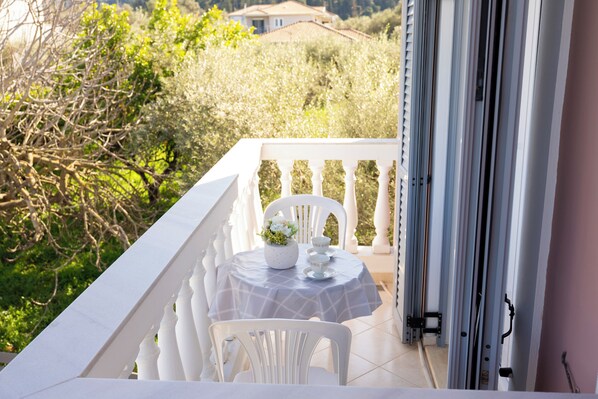 A balcony with a table set for two, white chairs, and a view of greenery and a building in the background. - Nikorina Apts | 4 (Apolpena)