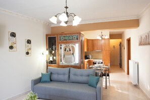 A living room with a sofa, a dining area with a table and chairs, a kitchen with cabinets and appliances, and a hallway leading to another room. - Nikorina Apts | 4 (Apolpena)