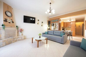 A living room with a sofa, coffee table, and a fireplace. There is a kitchen area with wooden cabinets and a dining table. - Nikorina Apts | 4 (Apolpena)