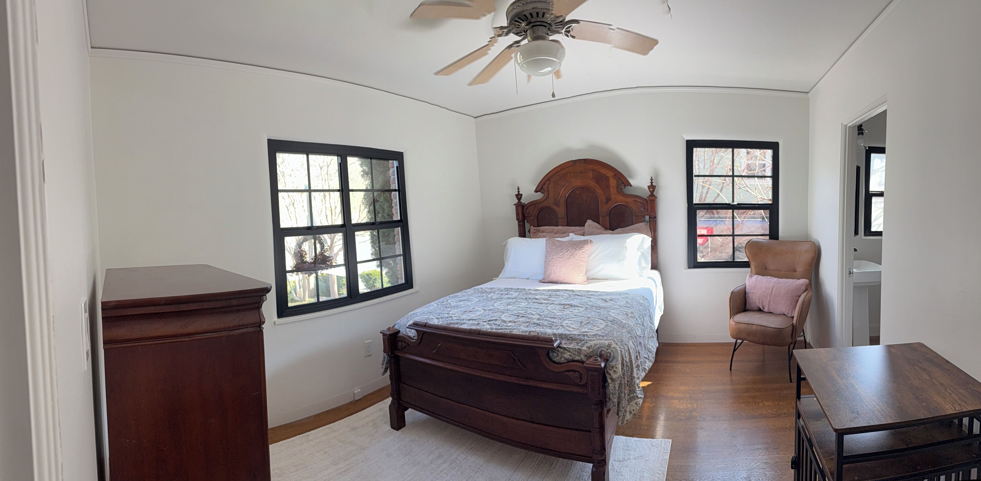 A bedroom with a wooden bed, two windows, a ceiling fan, a chair, and a small table.