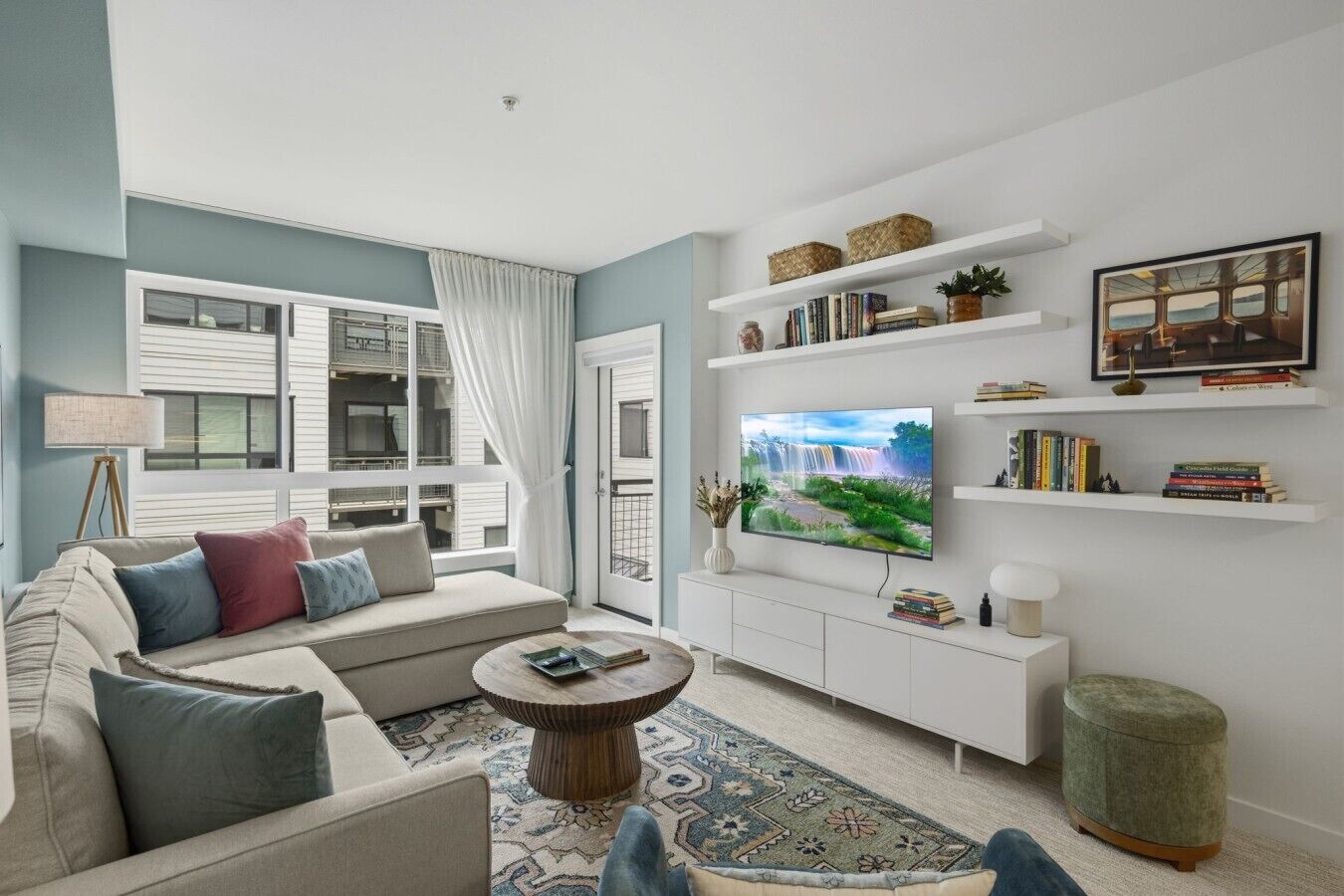A modern living room with a sofa, coffee table, TV, bookshelves, and a view of a building outside.