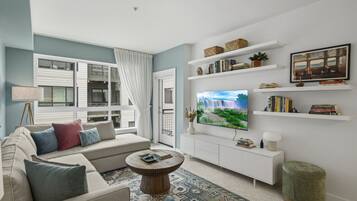 A modern living room with a sofa, coffee table, TV, bookshelves, and a view of a building outside.