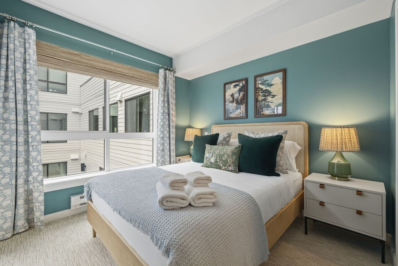 A bedroom with a large bed, two bedside tables, a window with curtains, and artwork on the wall.