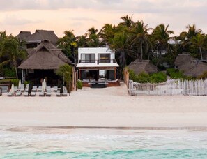 A beachfront property with a white building, thatched-roof structures, and a sandy beach. - Beach Home with Chef - Butler - Pool & Sun Deck (Tulum)
