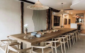 A long wooden dining table with chairs, a hanging light fixture, and a modern kitchen in the background. - Beach Home with Chef - Butler - Pool & Sun Deck (Tulum)