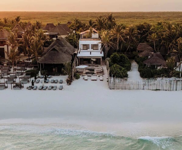 Une propriété située en bord de mer, comprenant un bâtiment principal, plusieurs constructions à toits de chaume et une plage de sable avec des chaises longues.