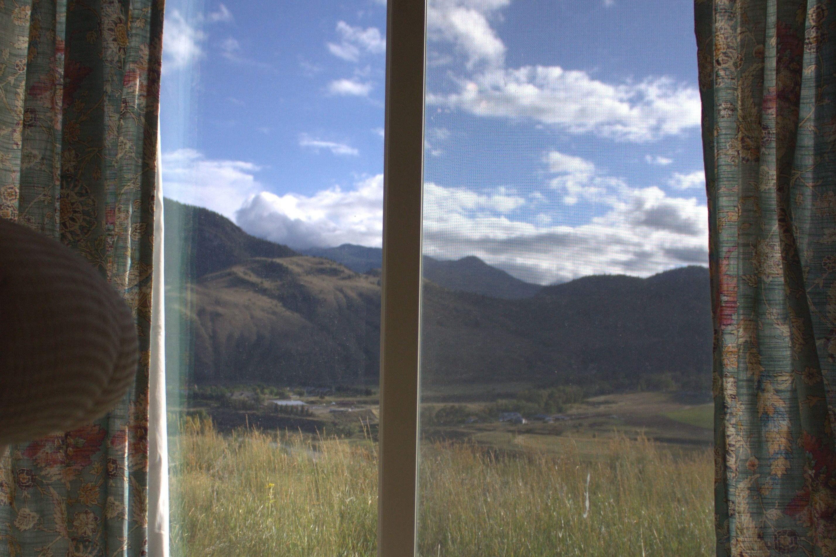 A window with a view of a mountainous landscape, curtains with a floral pattern, and a clear sky.