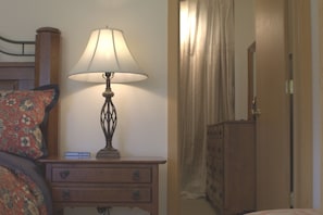 A bedside table with a lamp, a bed with a floral pillow, and a wooden dresser in the background. - A private apartment on six acres, with a view no other listing in Gardiner has. (Gardiner)