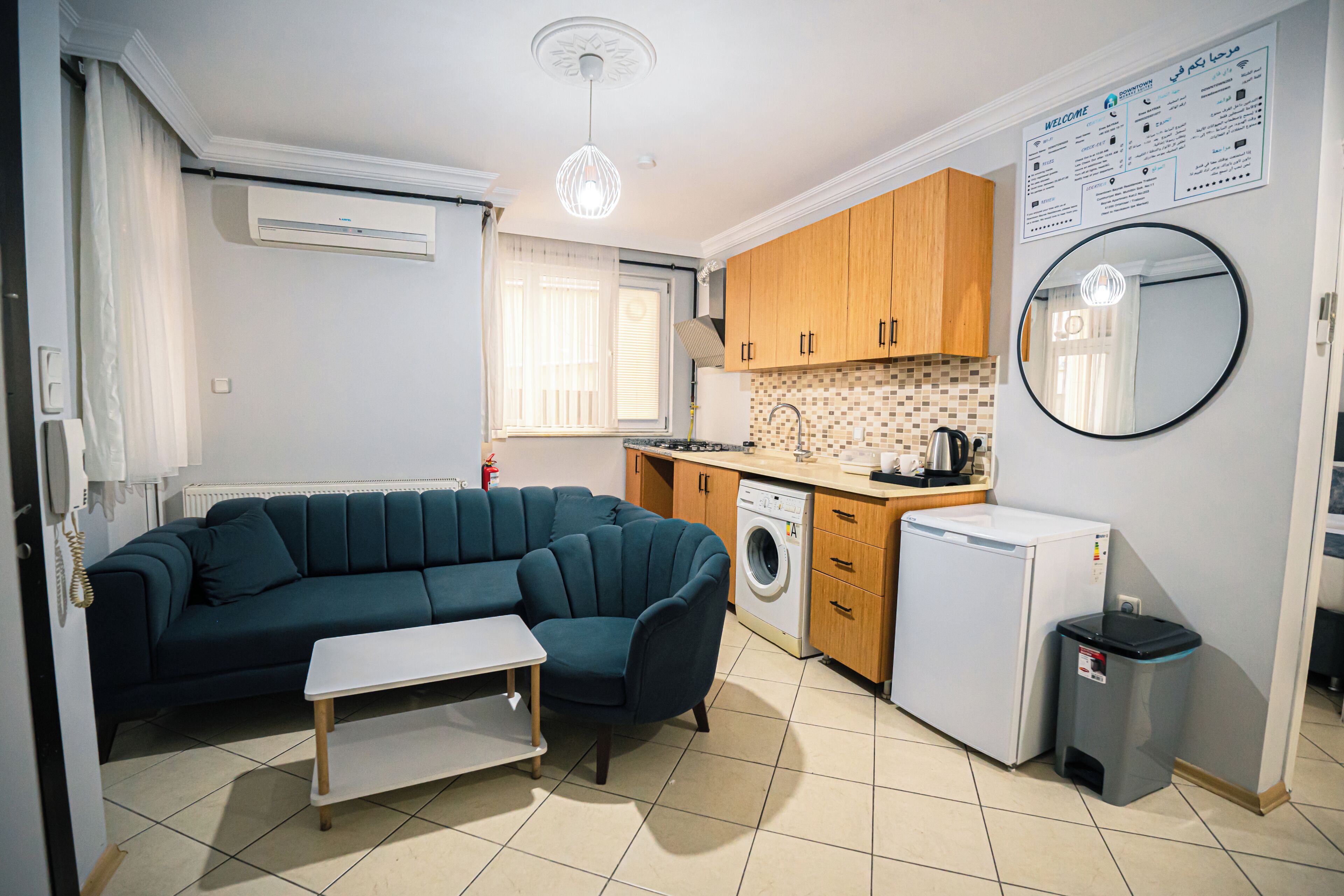 A compact living space with a sofa, a small table, a washing machine, a compact refrigerator, and a trash bin.