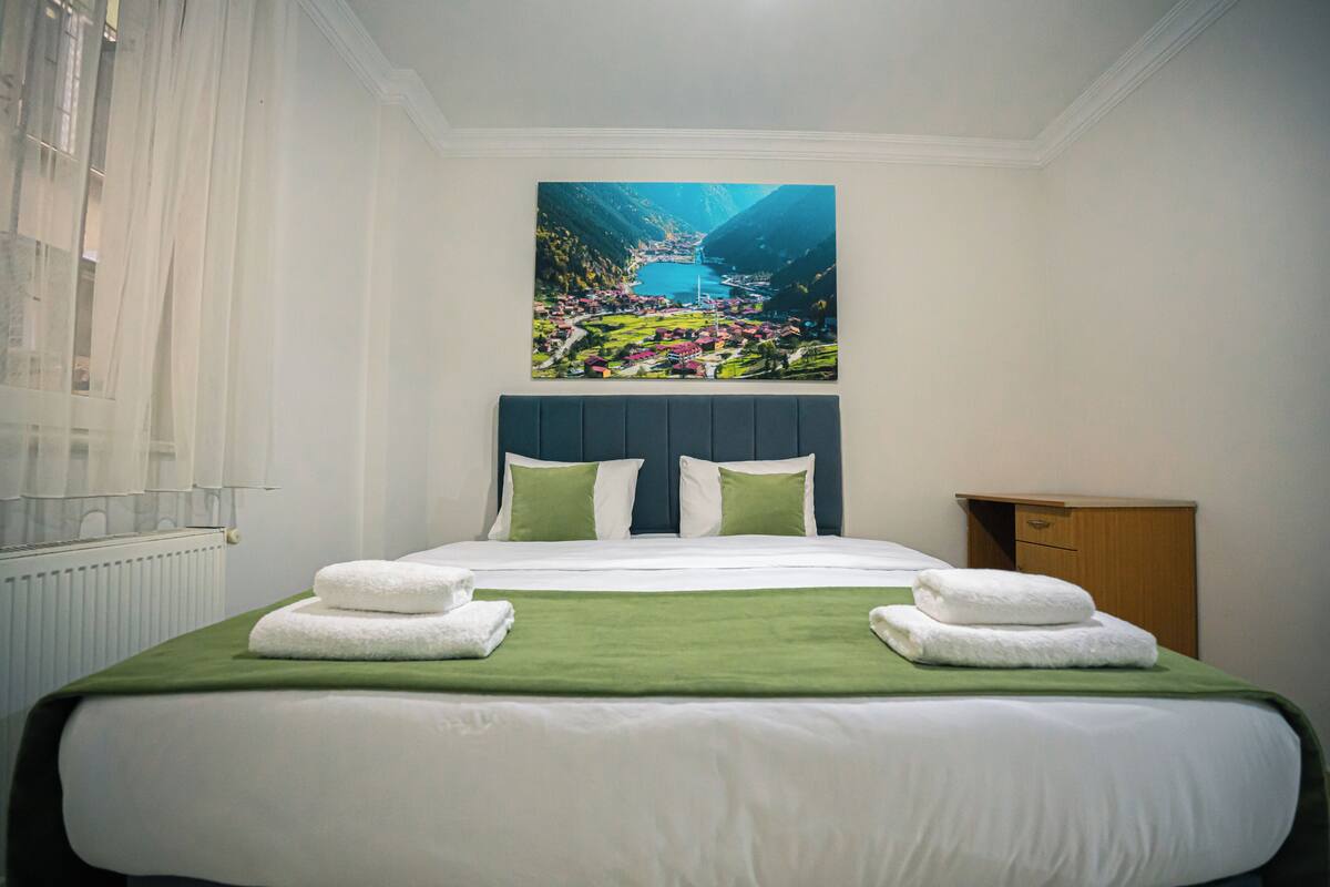 A hotel room with a large bed, a painting of a landscape, and a wooden nightstand.