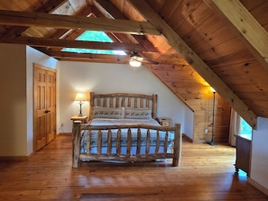 Un dormitorio con techo de madera inclinado, una cama con ropa de cama estampada, una mesita de noche con lámpara y una ventana con luz natural.