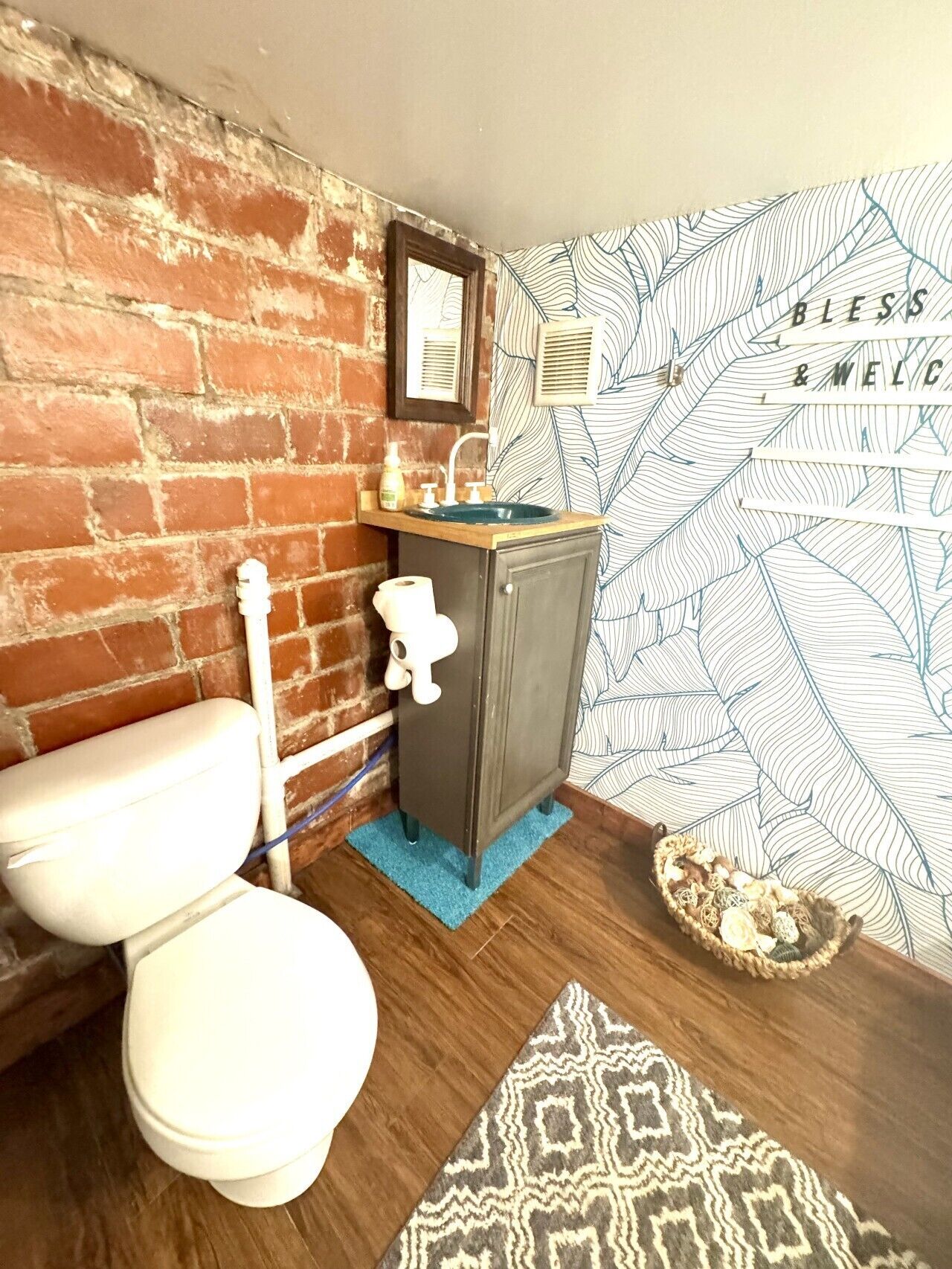 A bathroom with a brick wall, a wooden vanity with a sink, a toilet, and patterned wallpaper.
