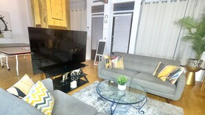 Sala de estar com sofá cinza, mesa de centro de vidro, televisão de tela plana e uma pequena planta.