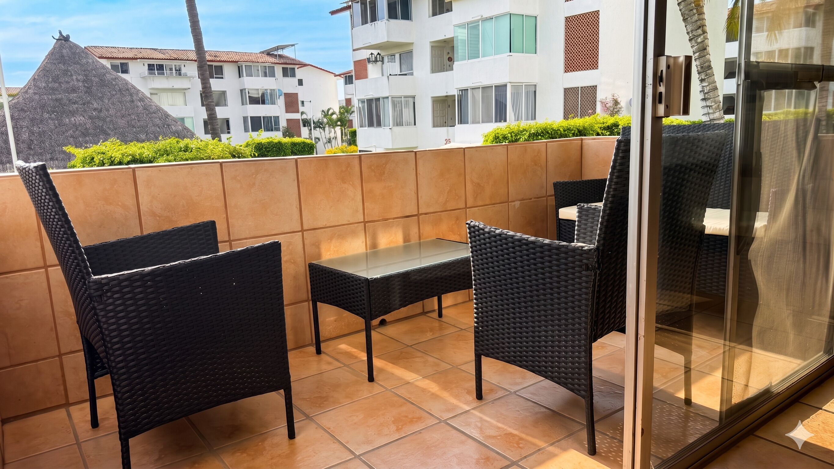 Un balcon avec deux fauteuils en osier et une petite table, une porte vitrée et une vue sur les bâtiments en arrière-plan.