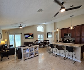 A spacious living area with a kitchen, dining area, and a TV displaying a golf course. - 4BR 3Bath Pool Home with Golf Course View, Quiet Cul De sac in Southern Dunes (Haines City)