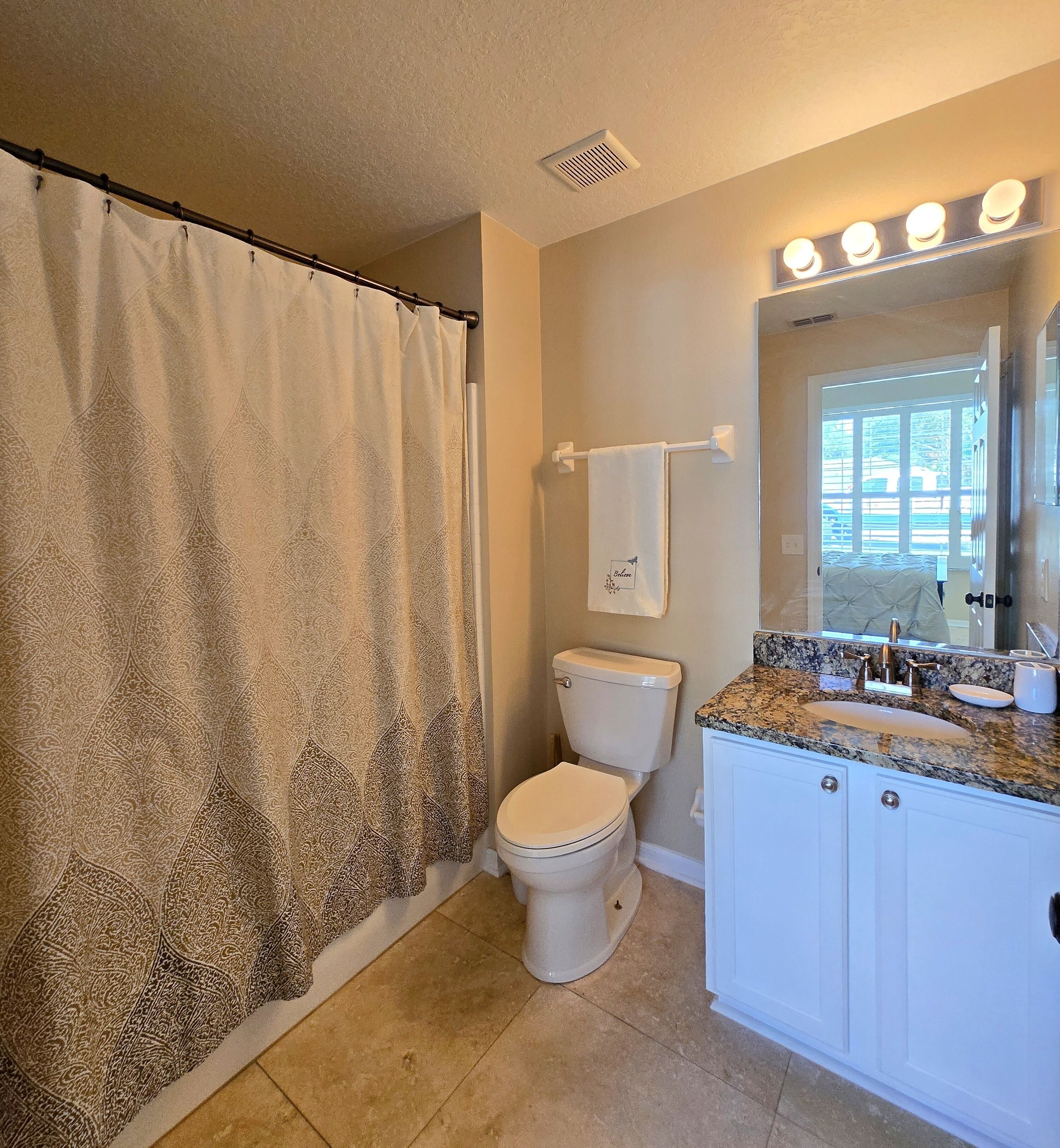 A bathroom with a patterned shower curtain, a toilet, a vanity with a sink, and a mirror with lights.