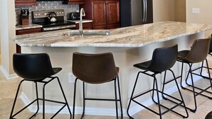 A kitchen with wooden cabinets, stainless steel appliances, a marble island, and bar stools. - 4BR 3Bath Pool Home with Golf Course View, Quiet Cul De sac in Southern Dunes (Haines City)