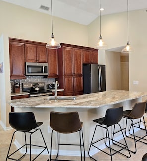 A kitchen with wooden cabinets, stainless steel appliances, a marble island, and bar stools. - 4BR 3Bath Pool Home with Golf Course View, Quiet Cul De sac in Southern Dunes (Haines City)