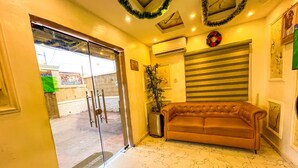 A hotel lobby with a glass door entrance, a brown leather sofa, a potted plant, an air conditioning unit, and a striped window blind. - Rosepearl Hotel And Suites (Lagos)