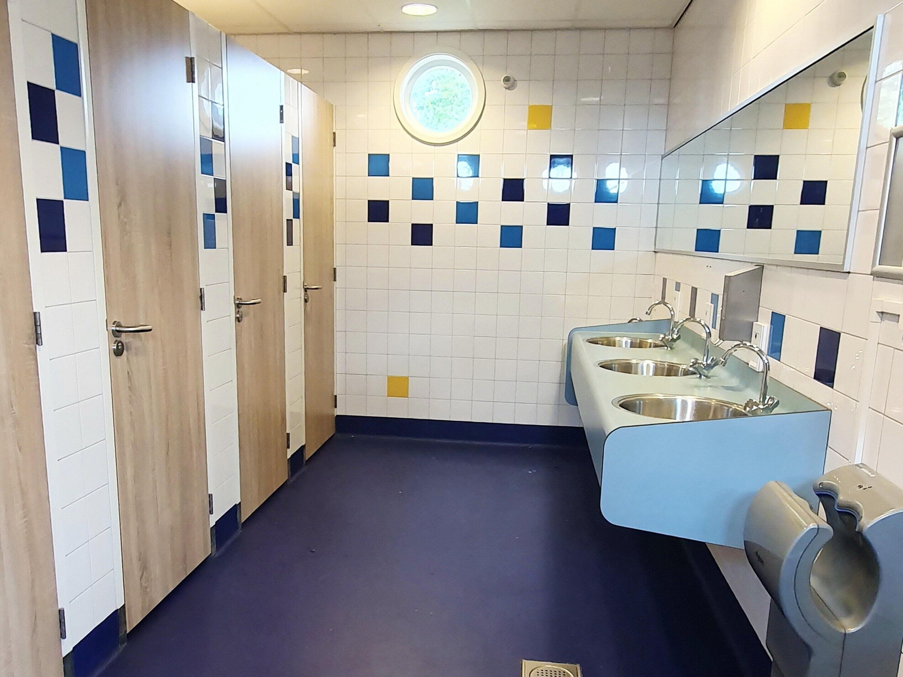 Un baño público con tres puertas de madera, suelo azul y pared de azulejos con cuadrados azules y amarillos.
