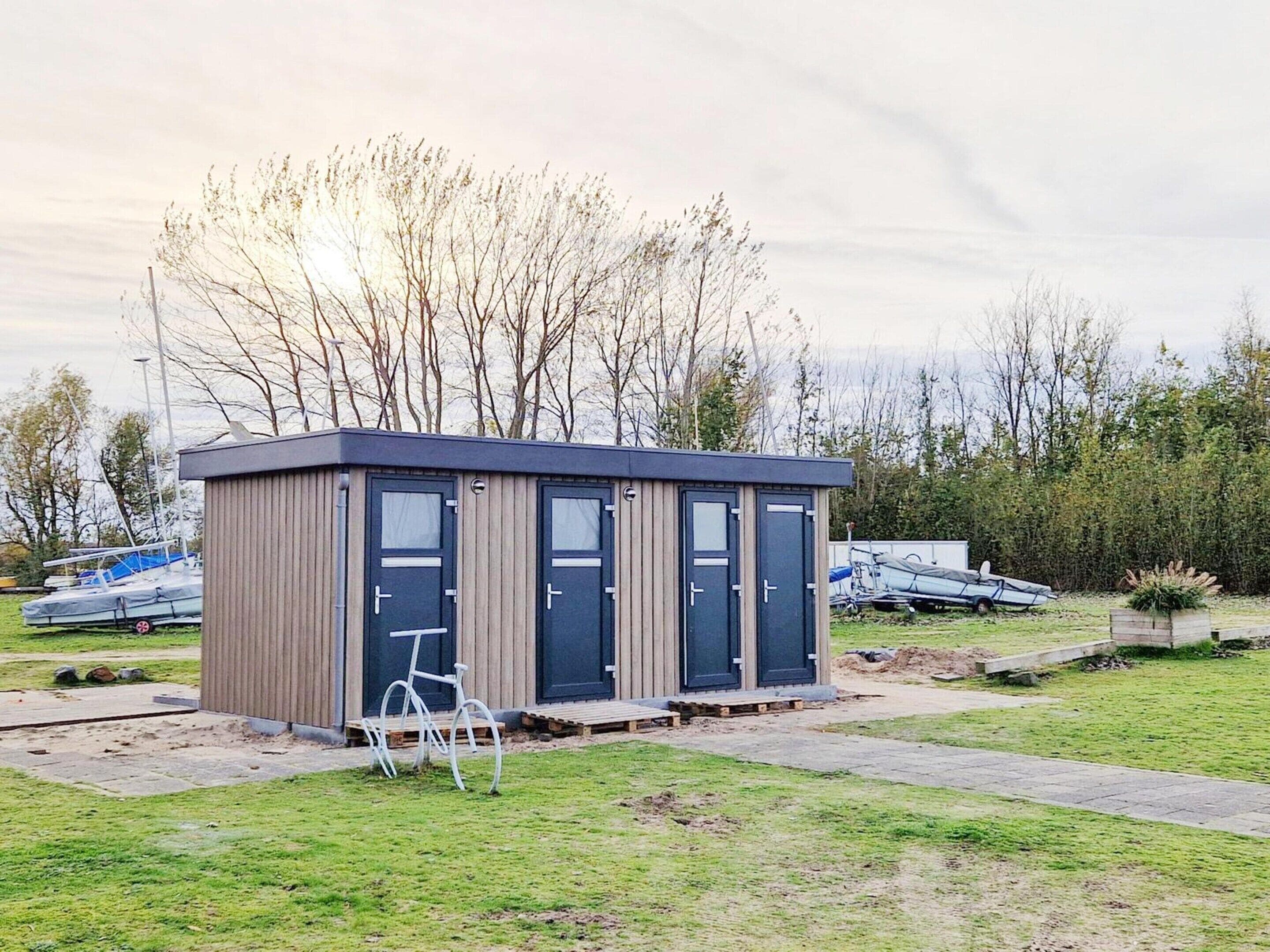 Een klein, modern gebouw met twee deuren en een raam, gelegen in een grasrijk gebied met bomen en boten op de achtergrond.