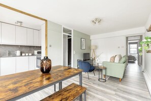 Ett modernt vardagsrum med ett matbord i trä, en soffa, ett litet sidobord och en köksdel med skåp och vitvaror.