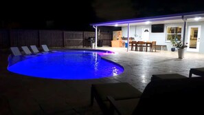 Ein Pool mit blauer Beleuchtung, eine überdachte Terrasse mit Essbereich und ein durch eine Hecke abgegrenzter Garten.