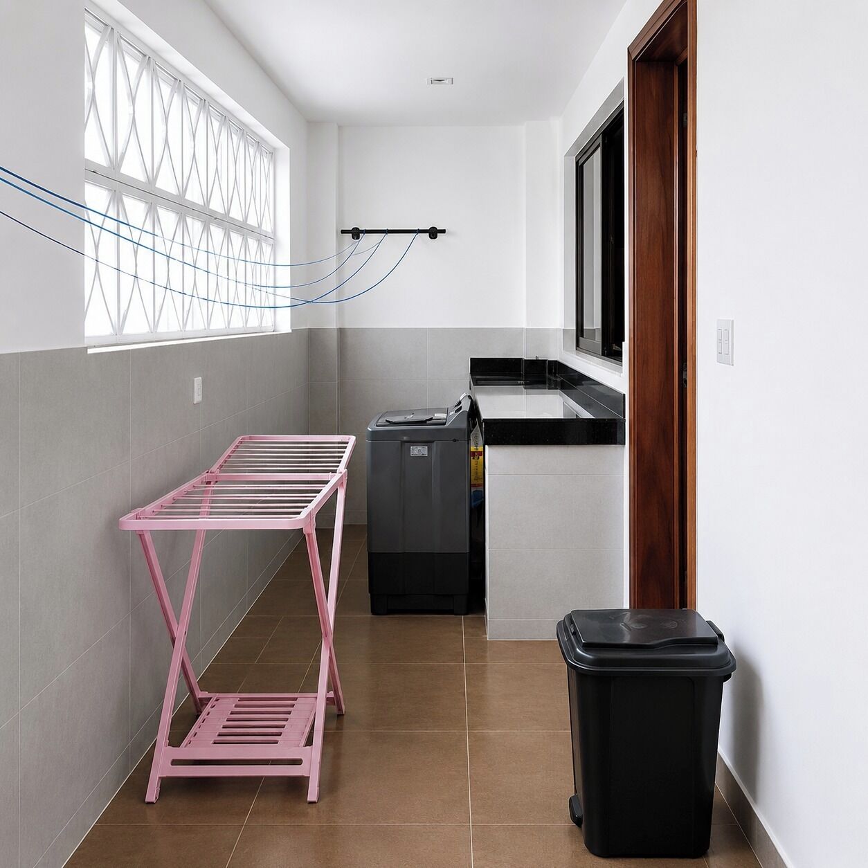 Un pasillo estrecho con un tendedero metálico rosa, un cubo de basura negro y una lavadora negra.