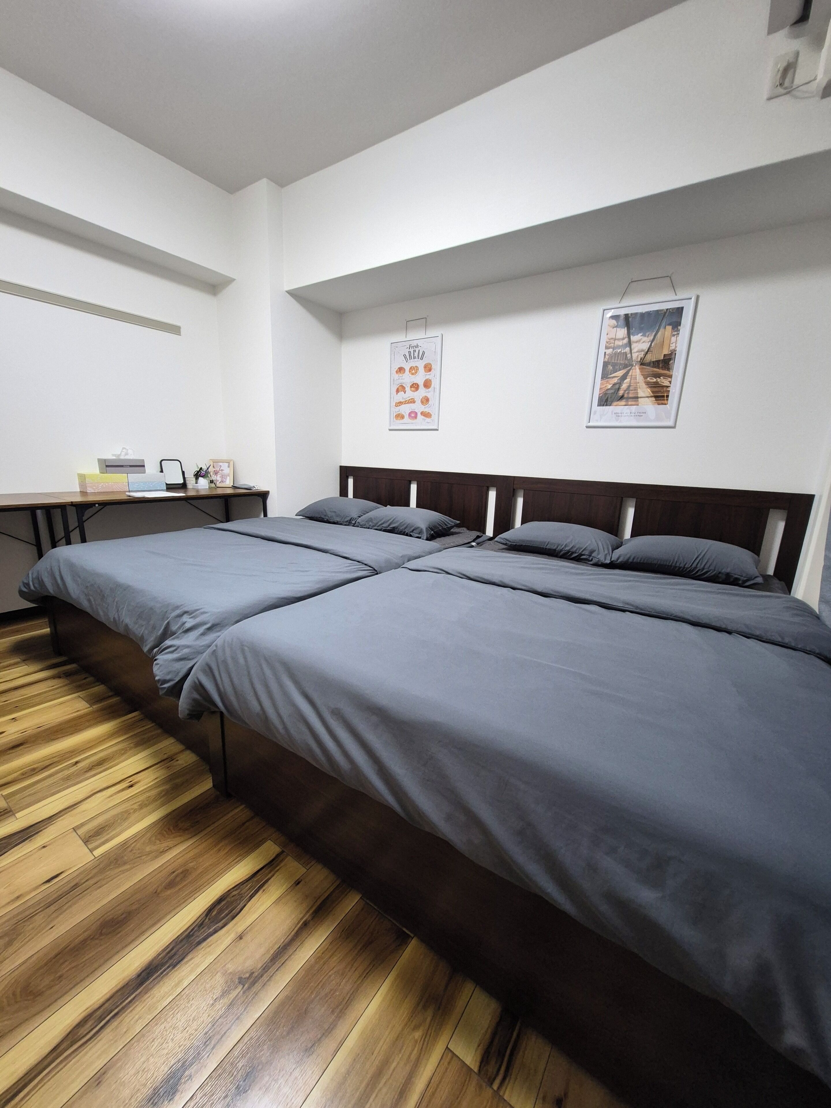 Ein Schlafzimmer mit Holzboden, einem Bett mit grauer Bettwäsche, einem Schreibtisch mit Stuhl und zwei gerahmten Bildern an der Wand.