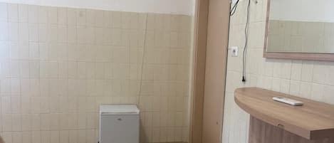 Un baño con piso de baldosas, un refrigerador blanco pequeño, una tocador de madera con espejo y un televisor montado en la pared.