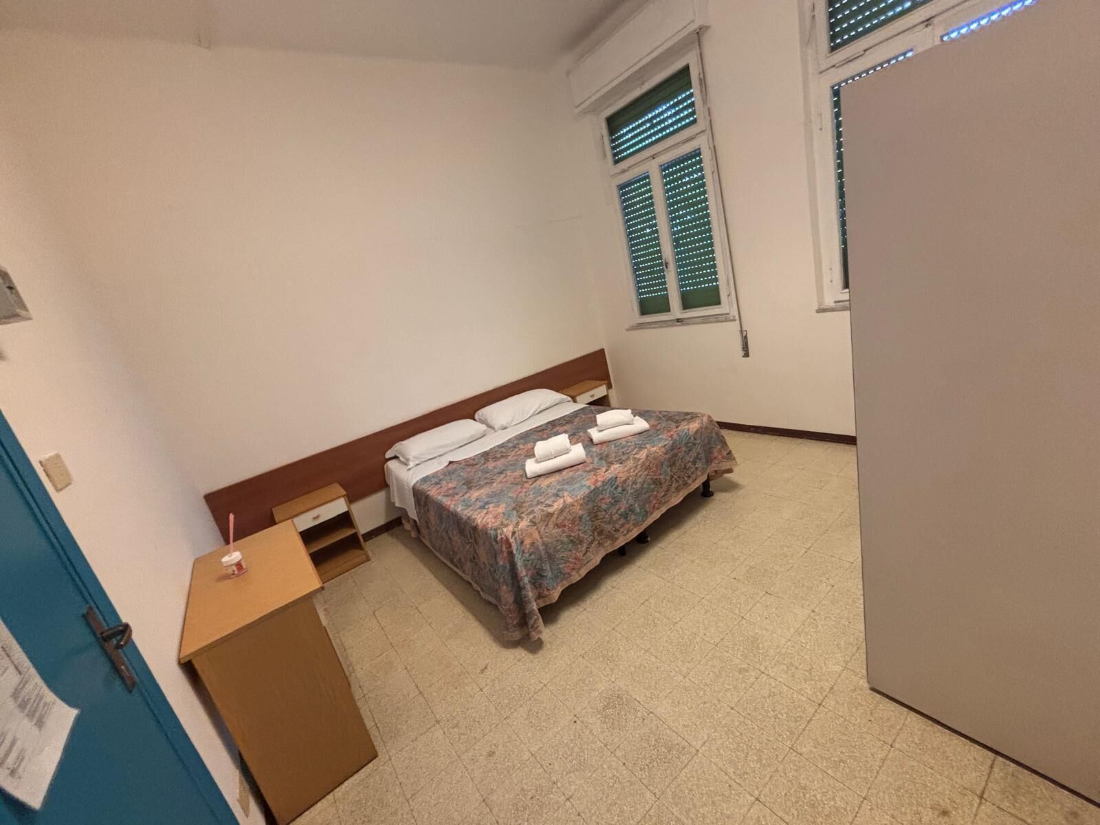 A bedroom with a bed, a desk, a window with blinds, and a door.