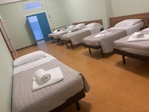 Habitación de hotel con cuatro camas, cada una con una toalla enrollada y una almohada. Se observa una puerta al lado izquierdo y una ventana encima.