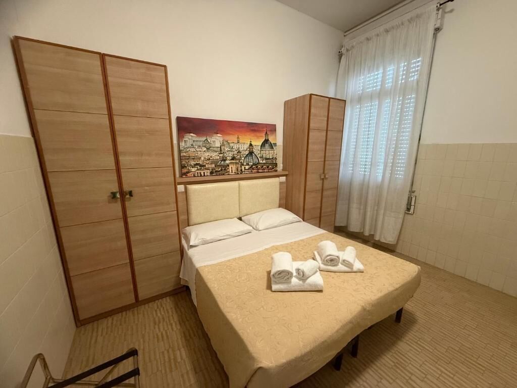 A hotel room with a bed, a painting of a cityscape, a wardrobe, and a window with curtains.