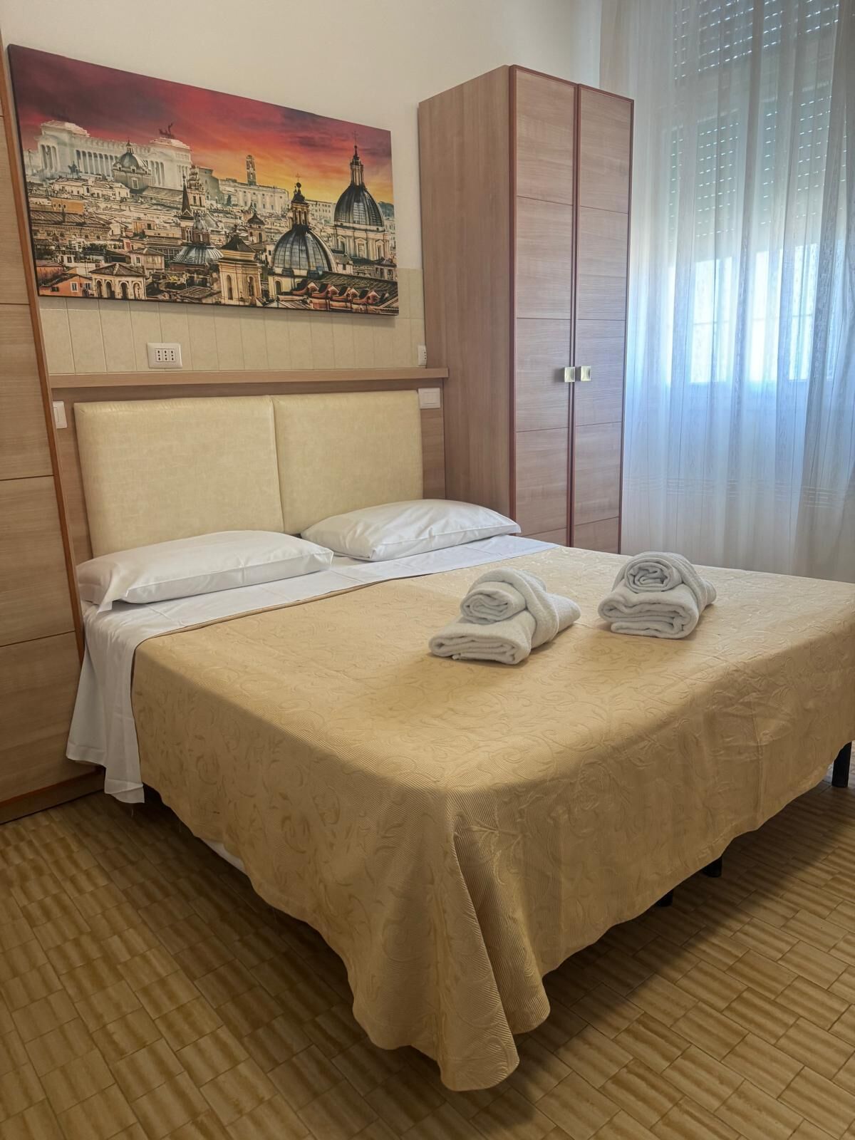 A hotel room with a bed, a headboard, a wardrobe, a painting, and a window with curtains.