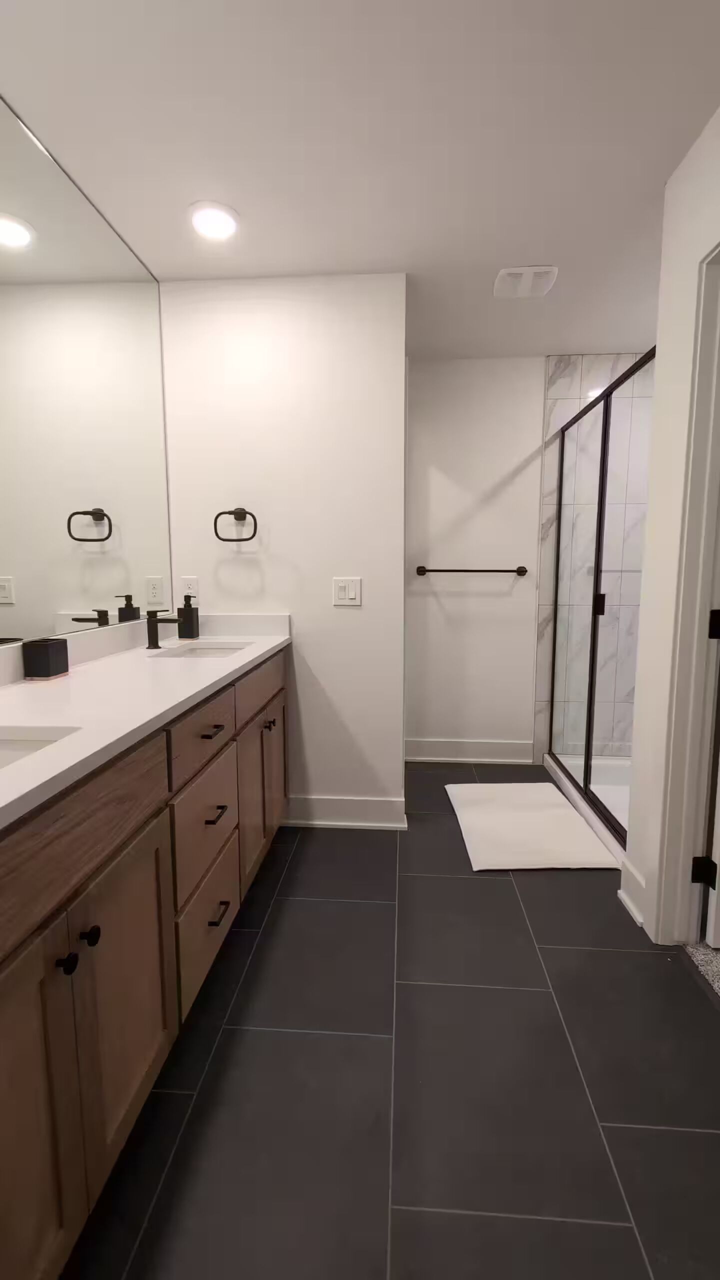 A modern bathroom with a large mirror, two sinks, wooden cabinets, a glass shower enclosure, and dark tiled flooring.