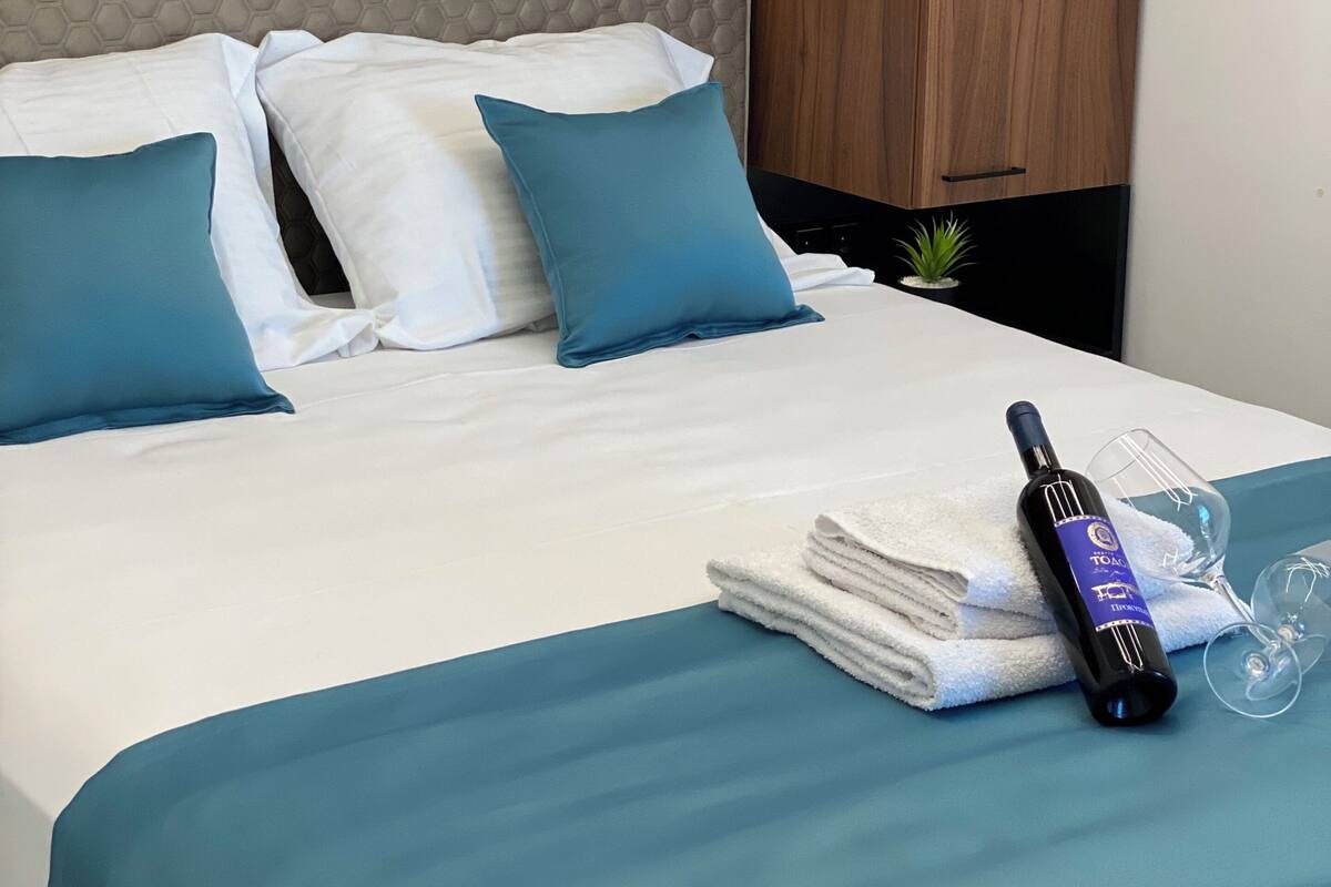 Una cama bien hecha con cabecera gris, almohadas blancas y turquesa, edredón turquesa y una botella de vino con vaso a un lado.