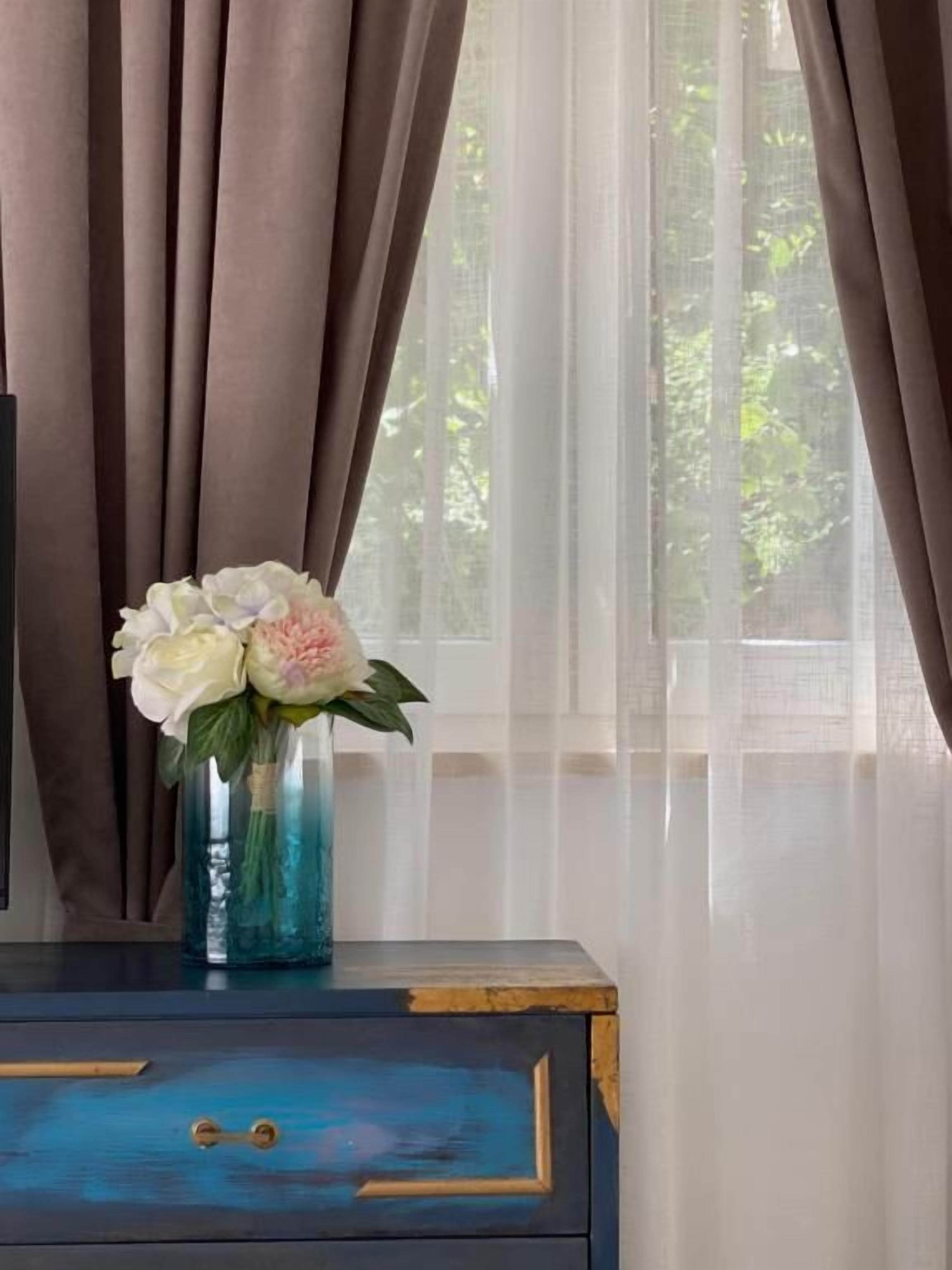 Una habitación con ventana, cortinas, un jarrón con flores y un aparador azul con cajones.