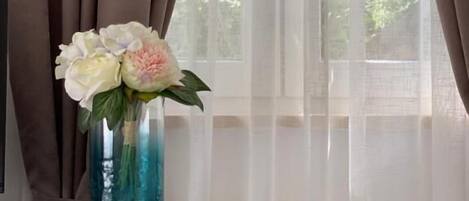 Una habitación con ventana, cortinas, un jarrón con flores y un aparador azul con cajones.