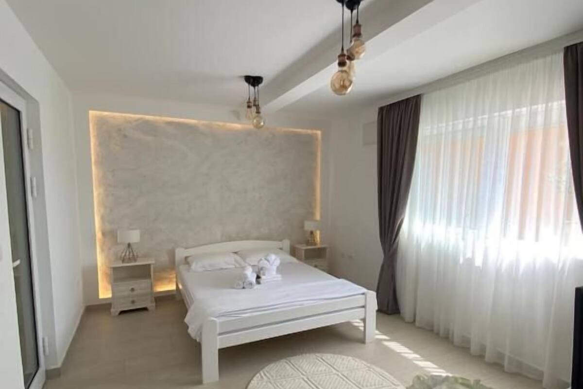 Un dormitorio moderno con una cama, mesitas de noche, una alfombra redonda y un jarrón con flores.
