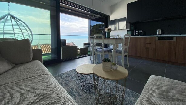 Un espacio habitable moderno con sofá, dos mesas redondas, una zona de cocina y un gran ventanal con vista al mar.